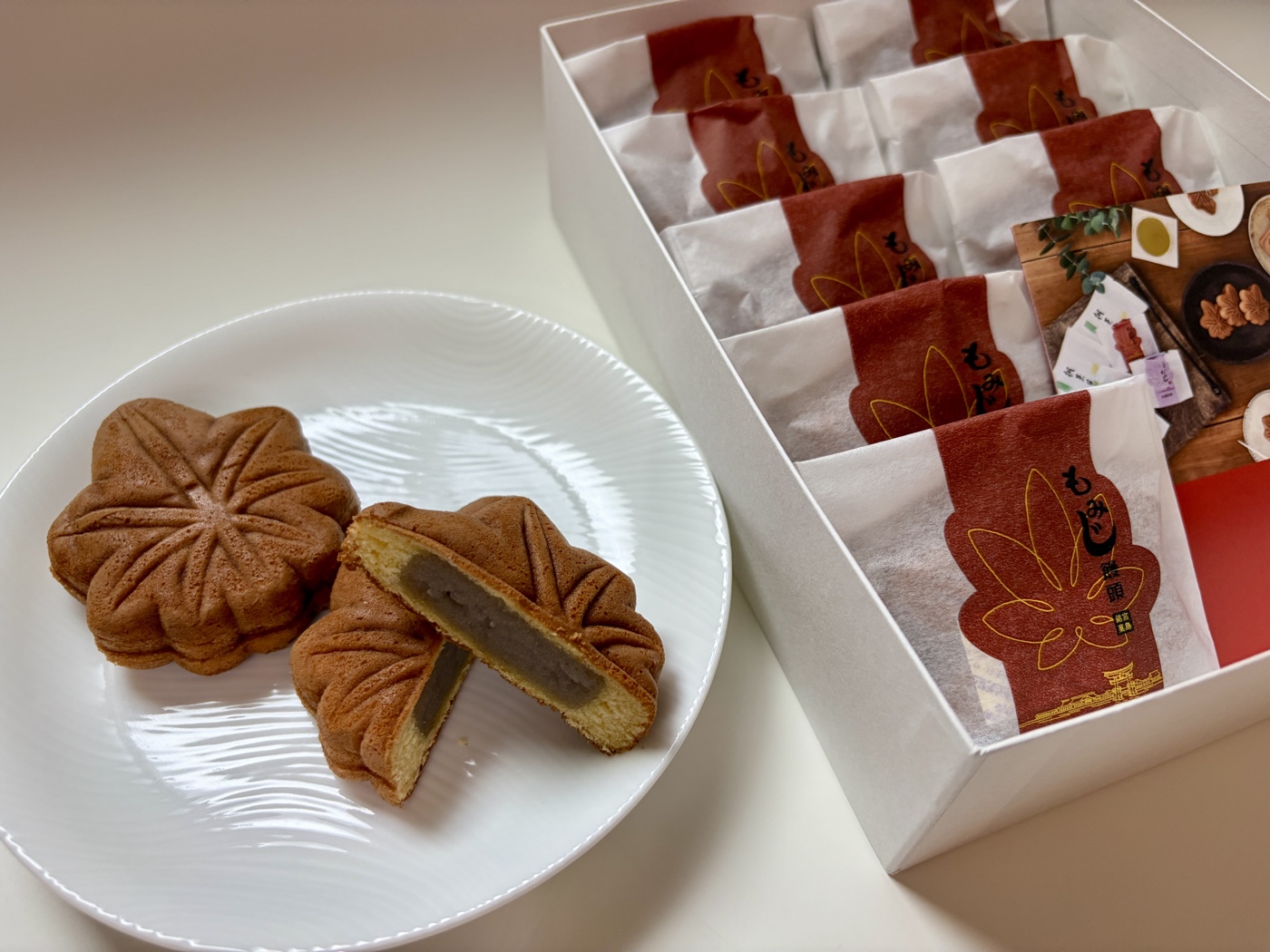 Momiji manju maple-leaf cakes — a gift box of individually wrapped pieces with two on a plate, one cut open to show the red bean filling — purchased and photographed by the Kasegeya editorial team (もみじ饅頭・編集部が実際に購入して撮影)